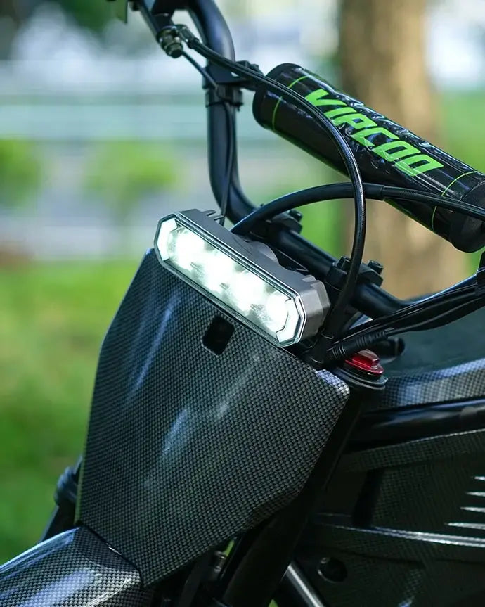 Close-up of the VIPCOO H3 Electric Dirt Bike by Vipcoo, featuring a bright LED headlight and handlebar pad with branding, set outdoors against blurred greenery.