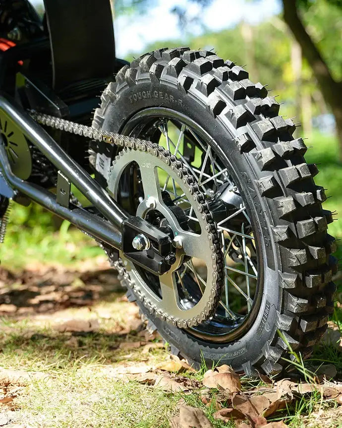 Close-up of the VIPCOO H3 Electric Dirt Bike by Vipcoo, showing its rugged off-road tire with deep treads, chain, and gears, parked on grass with dry leaves and sunlight streaming through trees in the background.