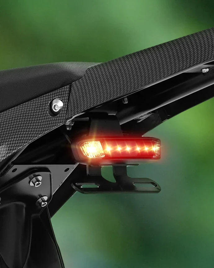 Close-up of the VIPCOO H3 Electric Dirt Bike’s rear shows an illuminated LED taillight and turn signal by Vipcoo, with a carbon fiber textured seat, set against a blurred green background.